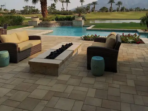 Paver pool deck with wicker sofas, a linear fire pit, and palm trees beside a resort-style pool